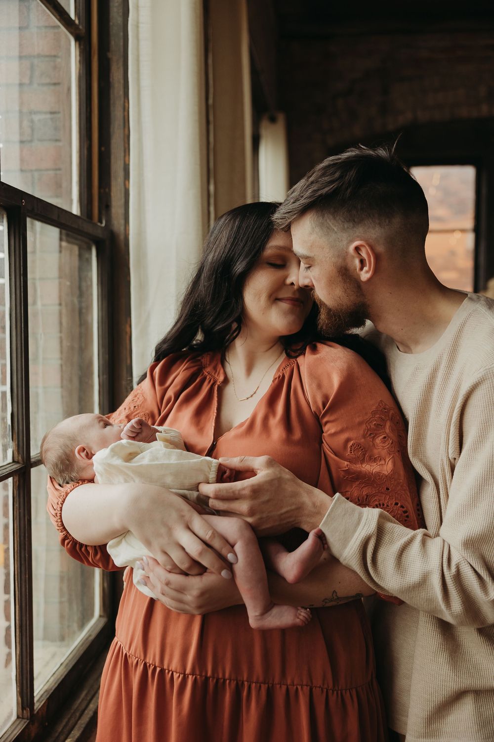 A couple in warm earthy tones shares an intimate moment with their newborn baby by a sunlit window.