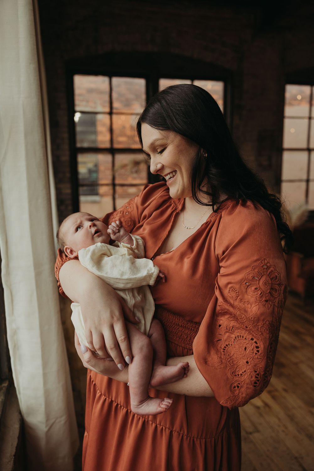 Parent in a rust-colored dress shares a joyful moment with their infant by a rustic window.