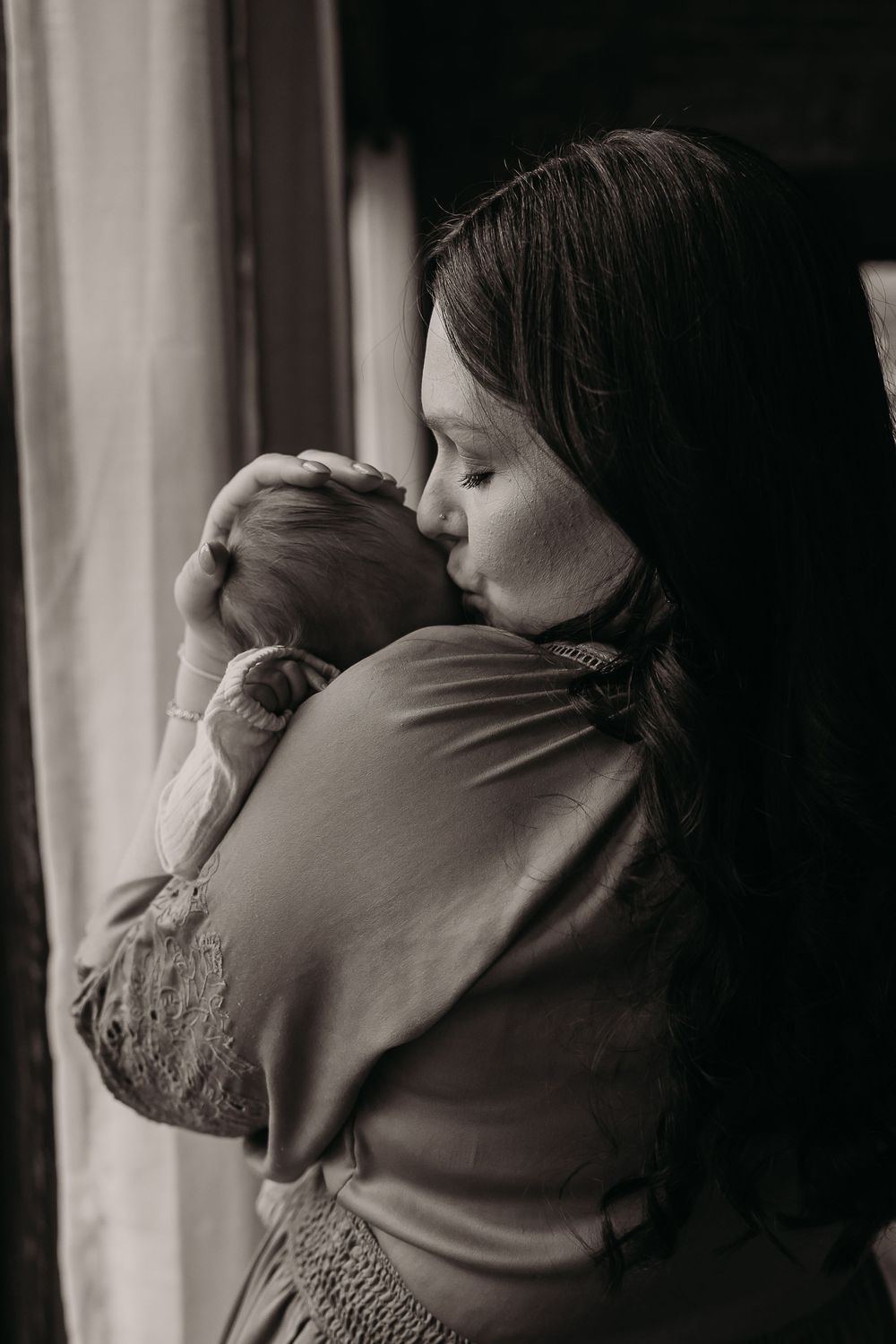 A tender moment between parent and newborn baby by a window in black and white photography.