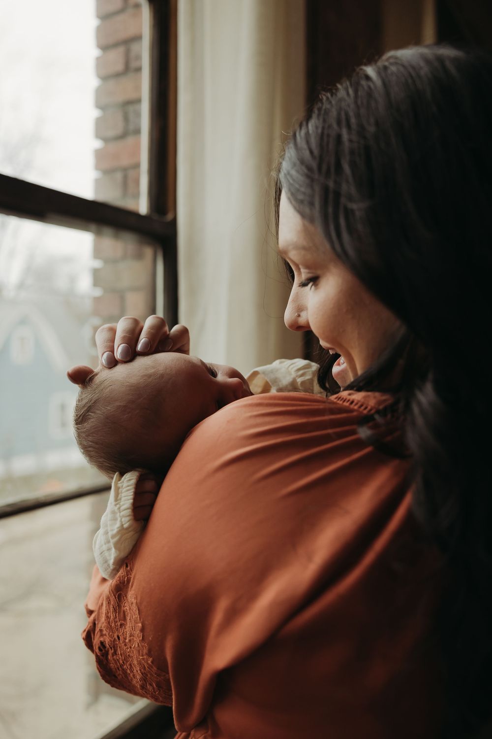 Person in orange sweater tenderly holding a sleeping infant while gazing lovingly near a window.