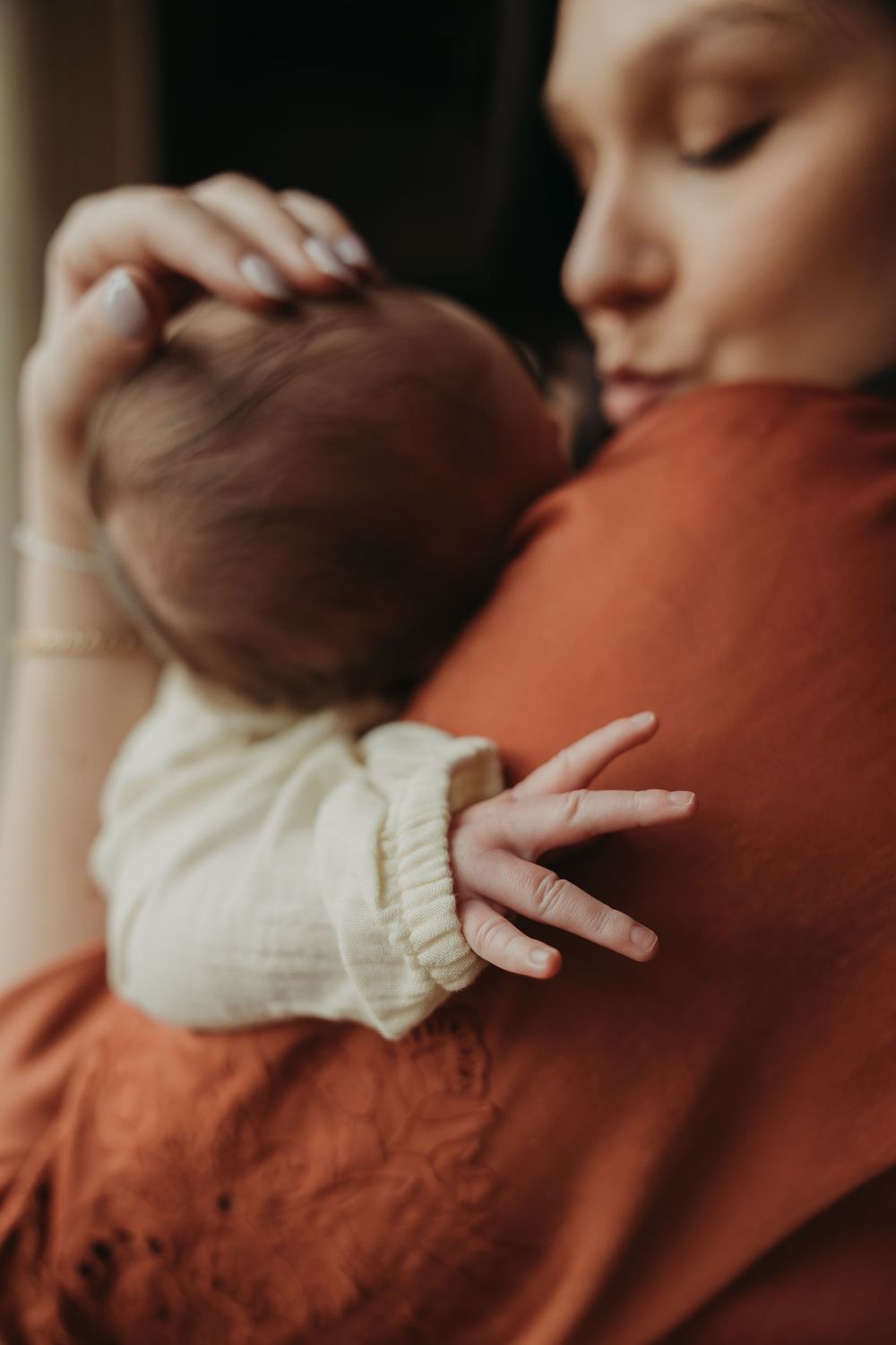 Close-up of parent in orange holding sleeping baby wearing cream sweater near window.