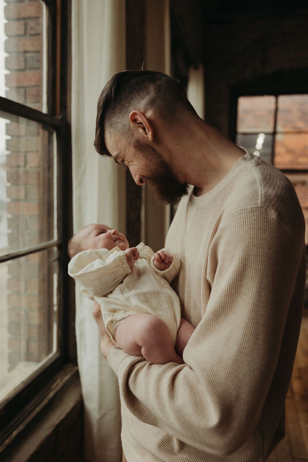 A tender moment by a window as a bearded person in a cream sweater cradles a small infant wrapped in white.
