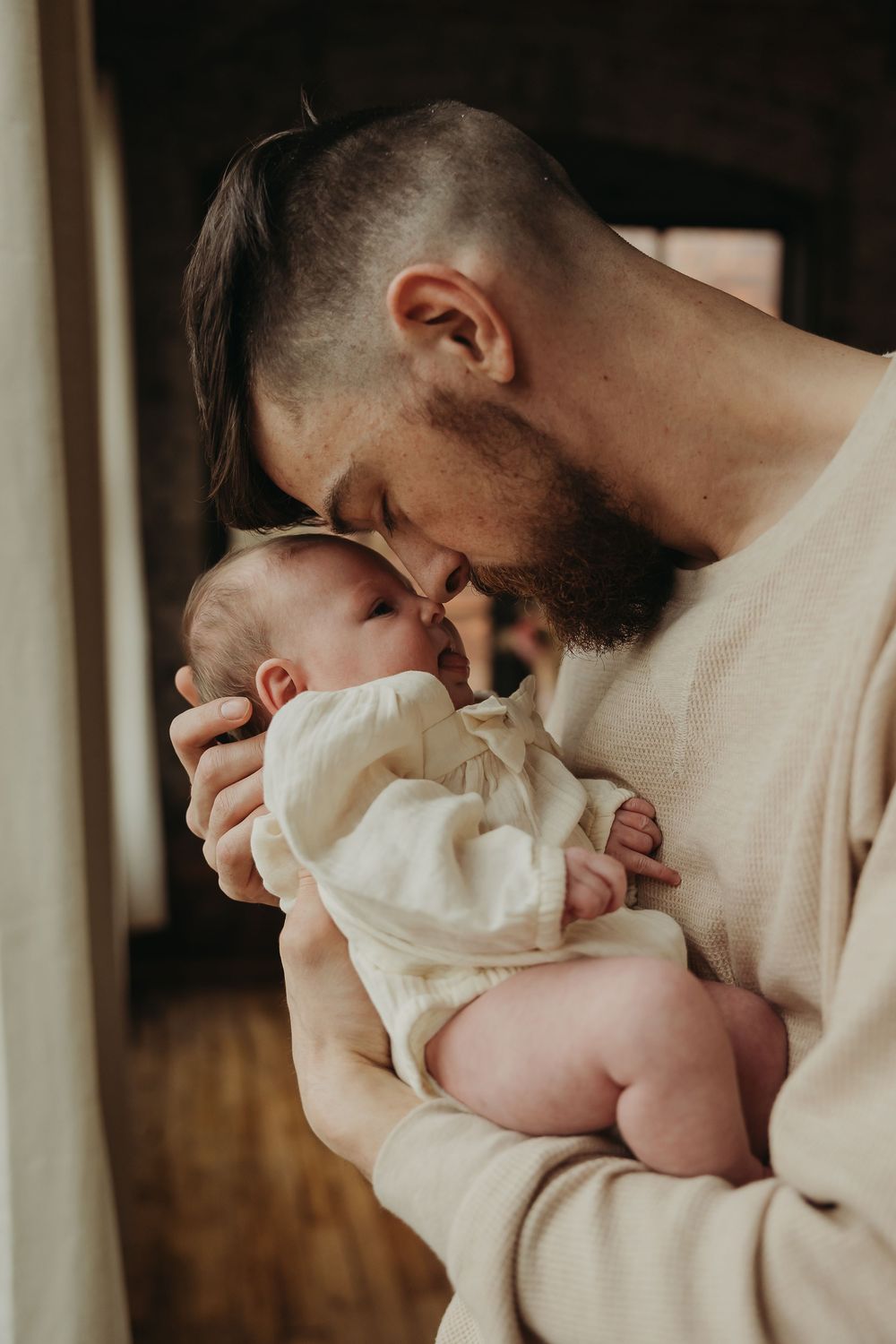 A tender moment between a parent and newborn baby in white clothing by a window.
