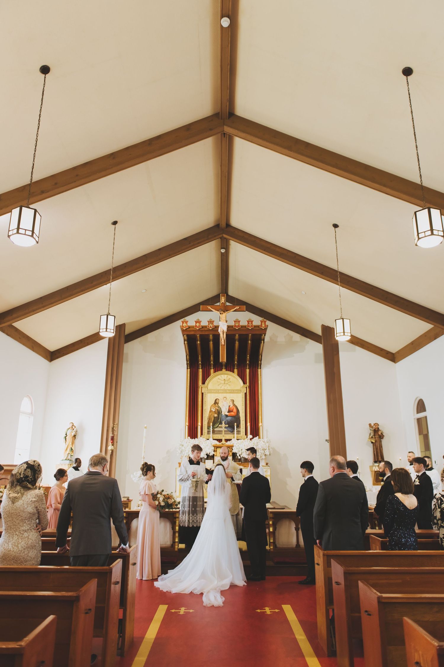 Traditional Latin Mass wedding ceremony takes place in Holy Family Parish with wooden beams and pendant lights under vaulted ceiling.