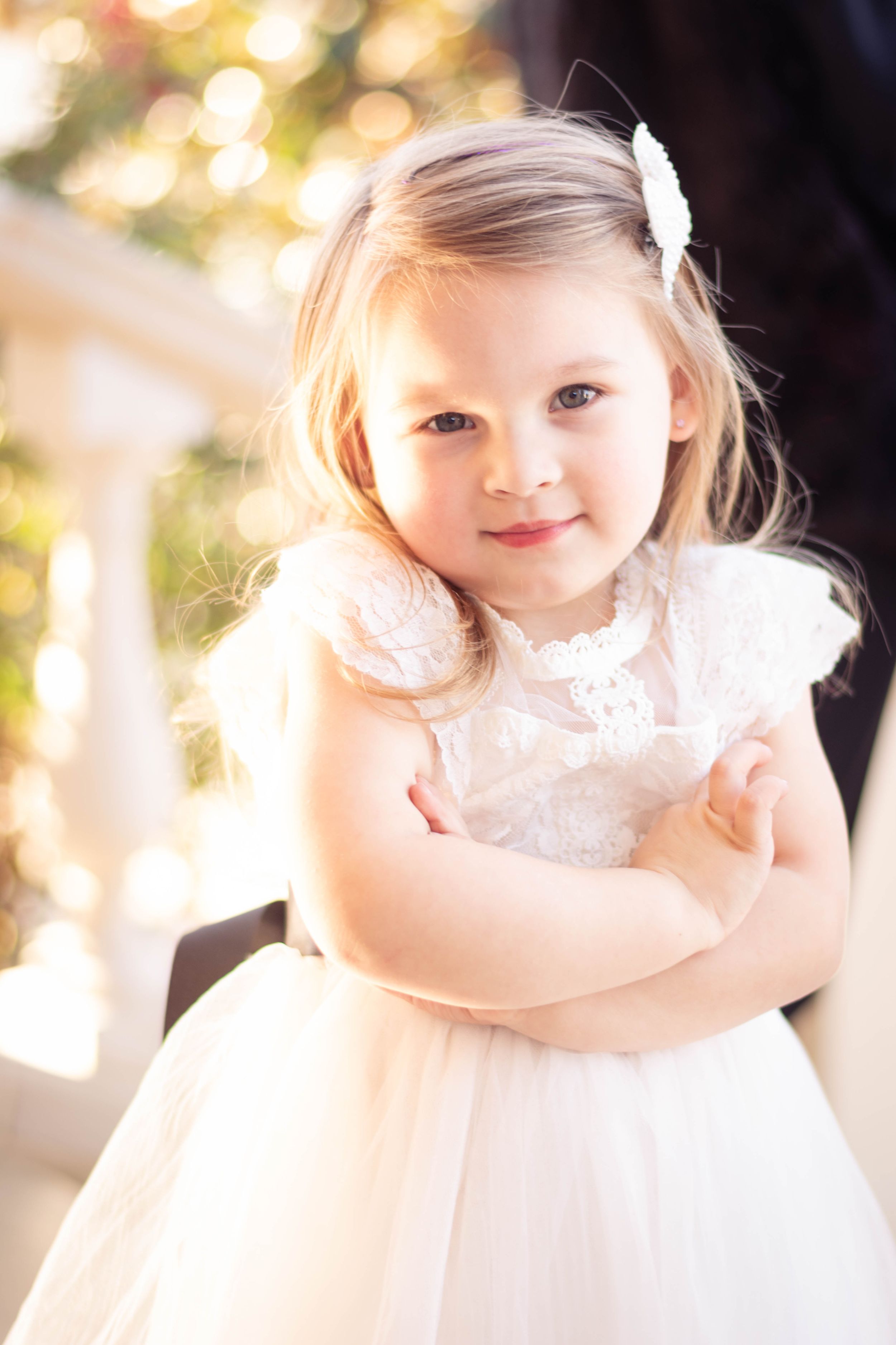 Young child wearing a white lace dress poses in soft natural light.