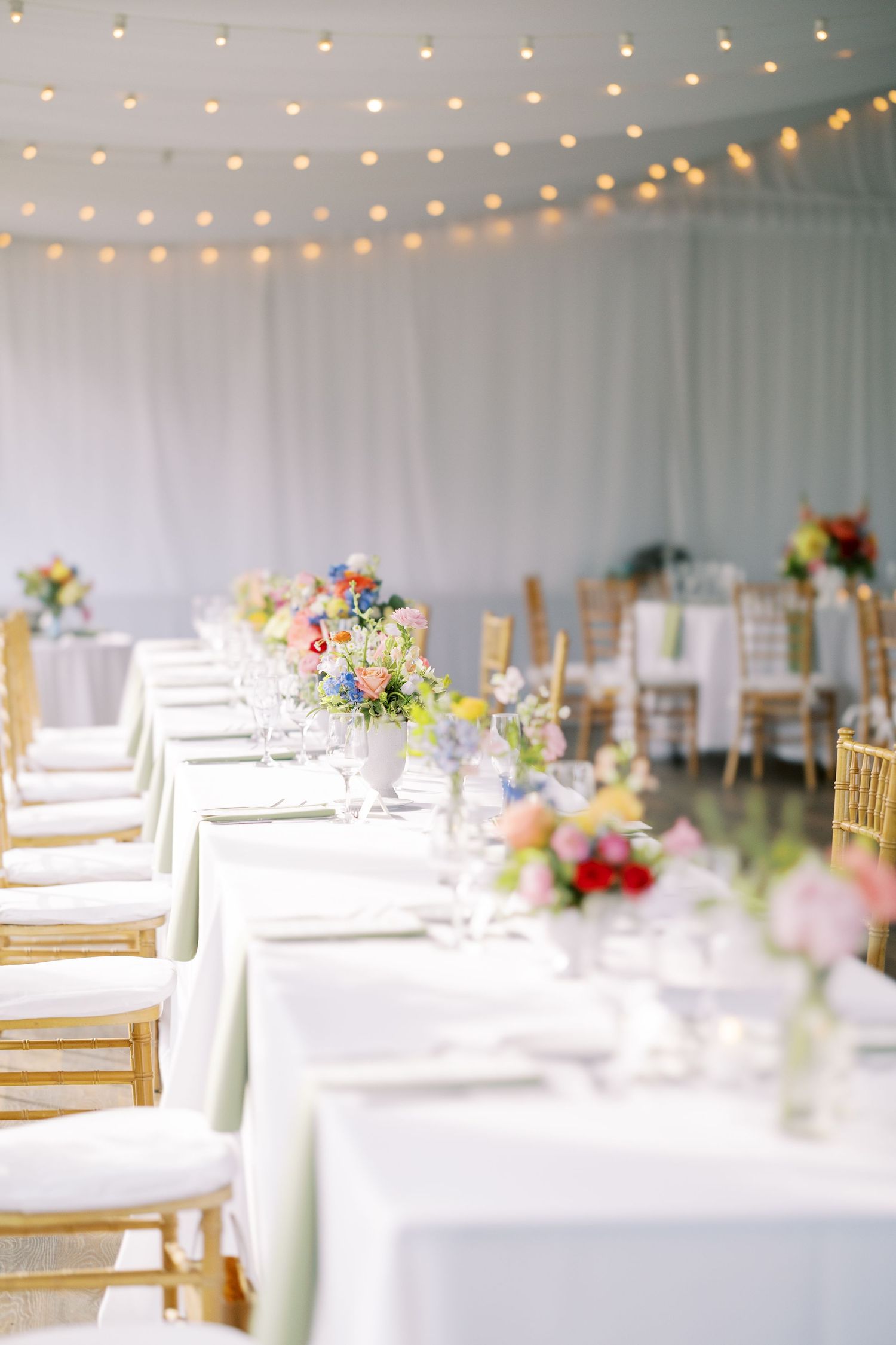 White banquet tables with gold chairs and string lights create an elegant wedding reception setting.