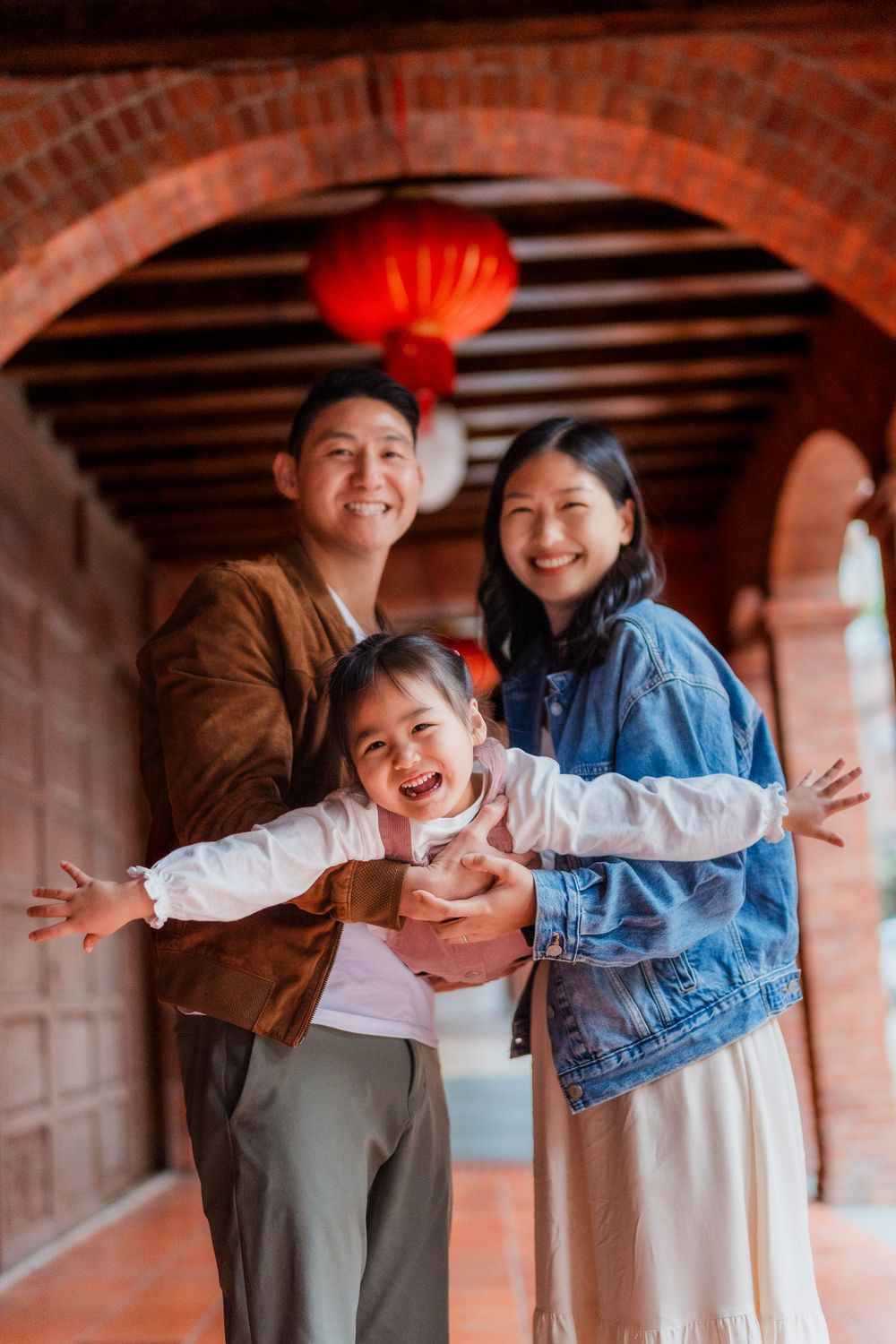 Parents and a young girl enjoying playful moments in a brick archway corridor with traditional red Chinese lanterns.