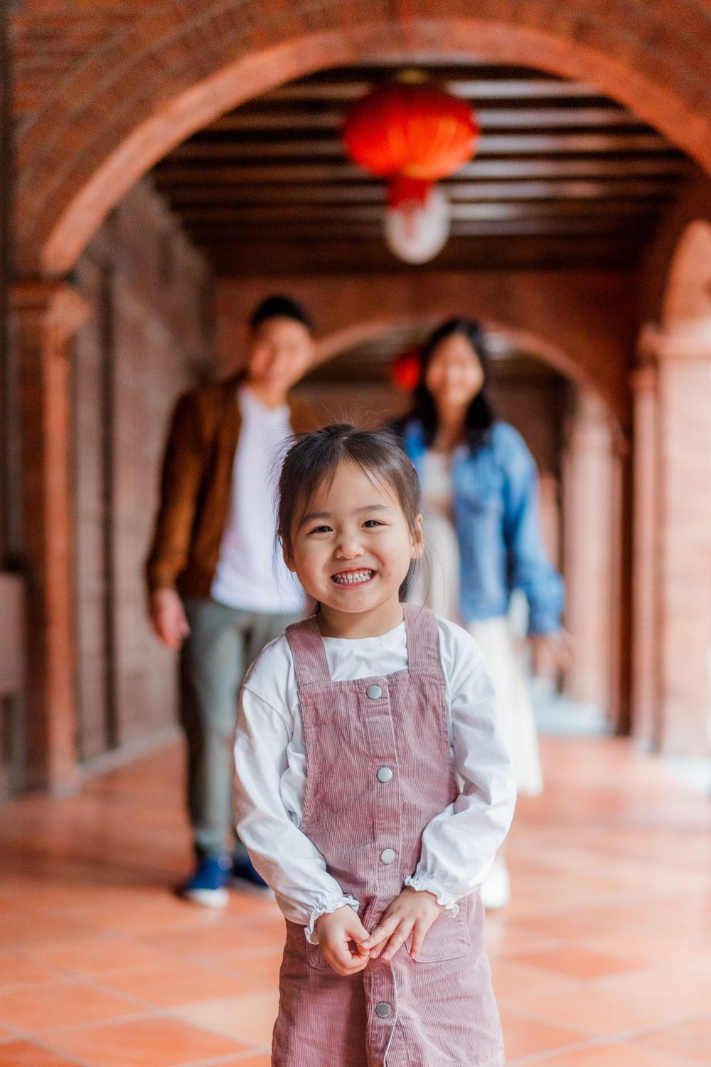 A smiling child in pink overalls and her parents at a distance, standing in a arched brick corridor with traditional Asian style lanterns.