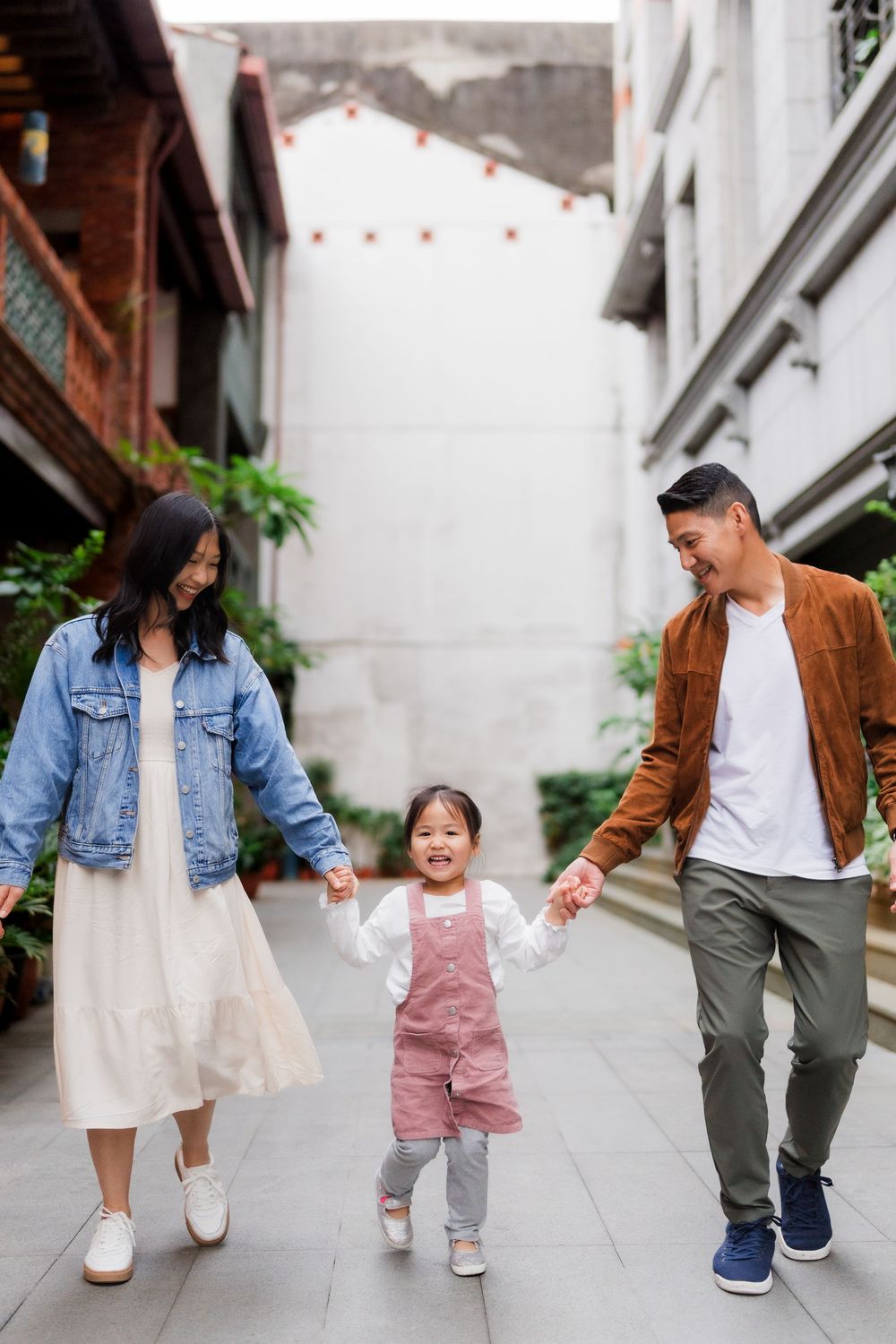 An aesthetic shot showing a child in pink overalls holding hands and walking between her parents in a softly light outdoor corridor.