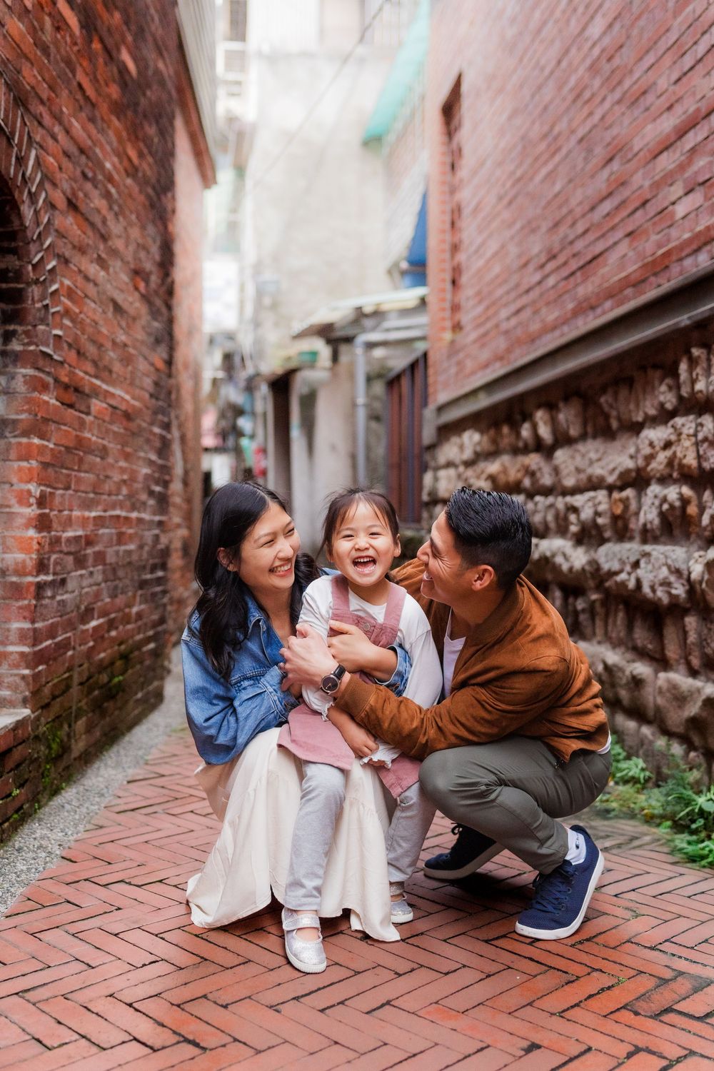 Joyful family moment captured on brick pavement in a narrow alleyway setting as two parents kneel down on either side of their young child.