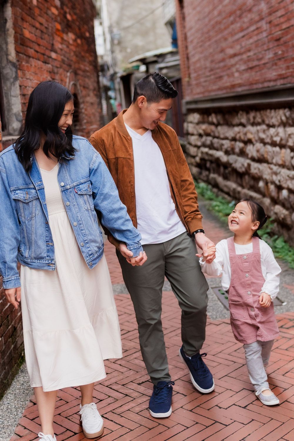 Family members walk together hand-in-hand smiling at one another in a brick alley wearing coordinated casual outfits.