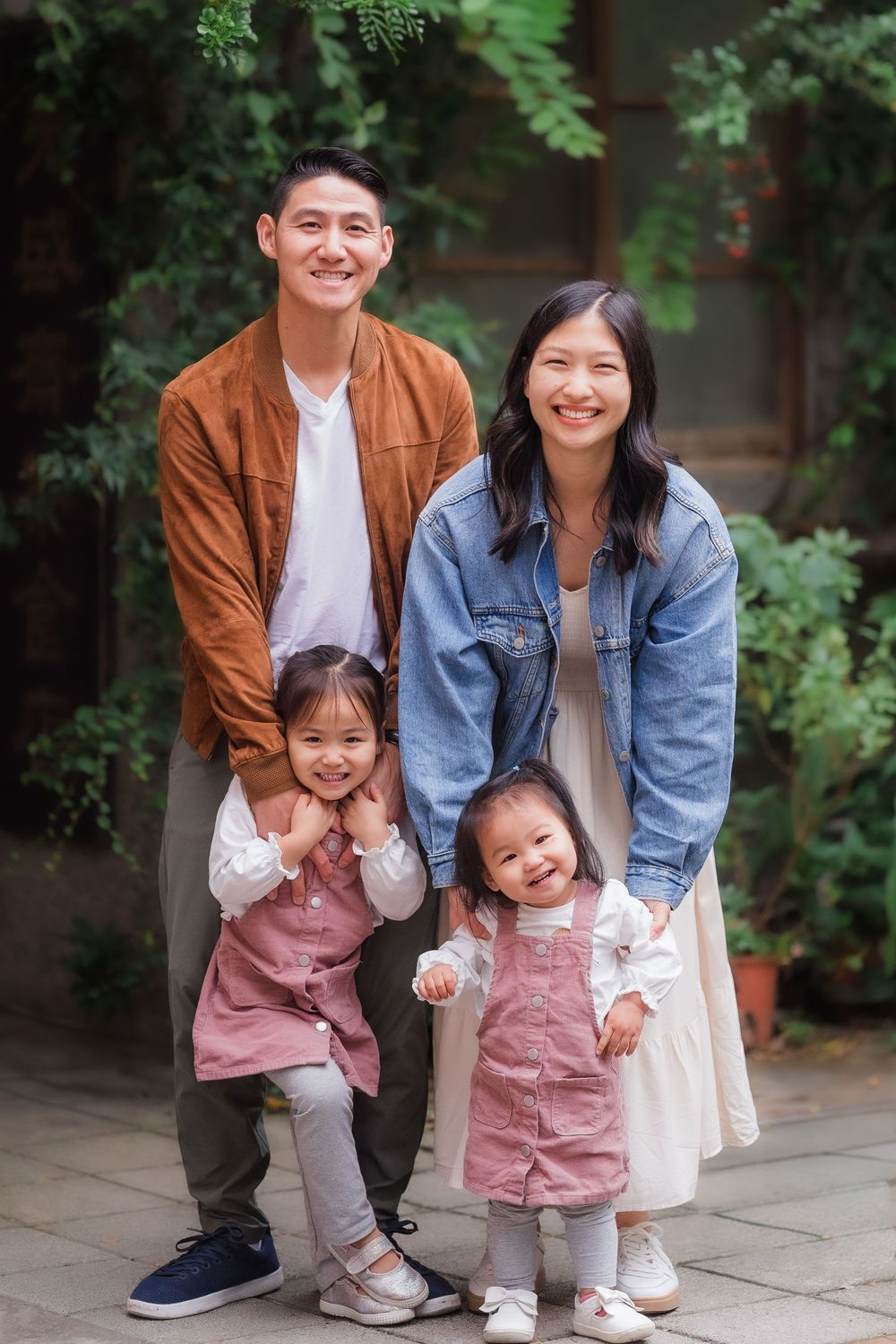 A casual family portrait of a happy family with two small children in coordinated pink overall dresses with green foliage in the background.