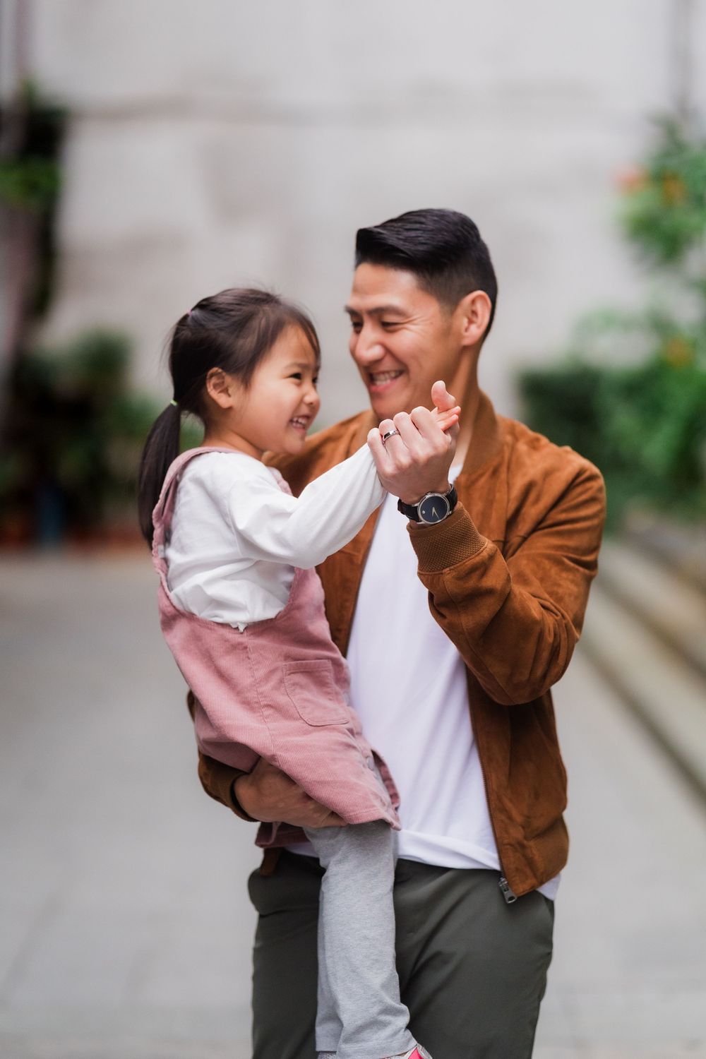 Playful sequence of a father holding his young daughter wearing pink overalls in an outdoor urban setting.