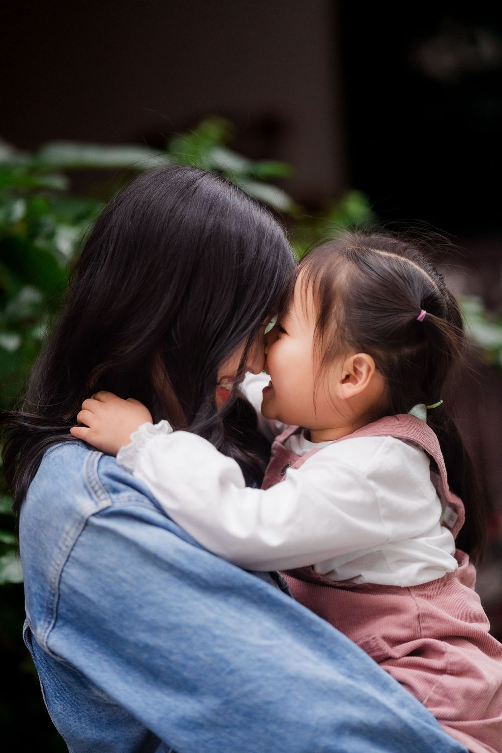 An intimate moment captured between a mother and daughter touching noses wearing denim and white clothing.