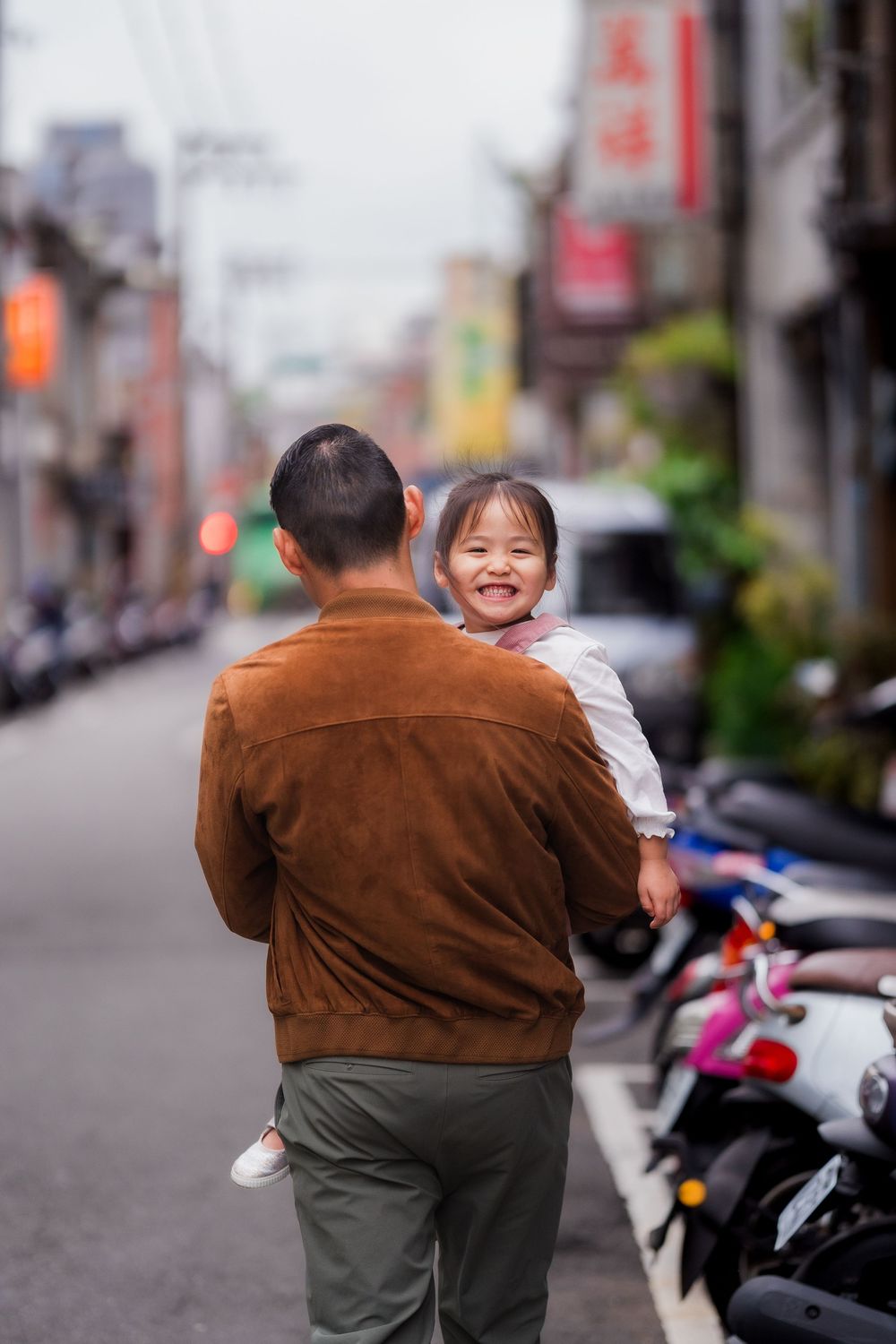 A man in a brown jacket carries a smiling small child down a city street lined with parked motorcycles.