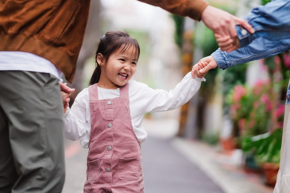A child in a pink overall dress smiles and holds hands with their parents while walking down a small street lined with greenery.