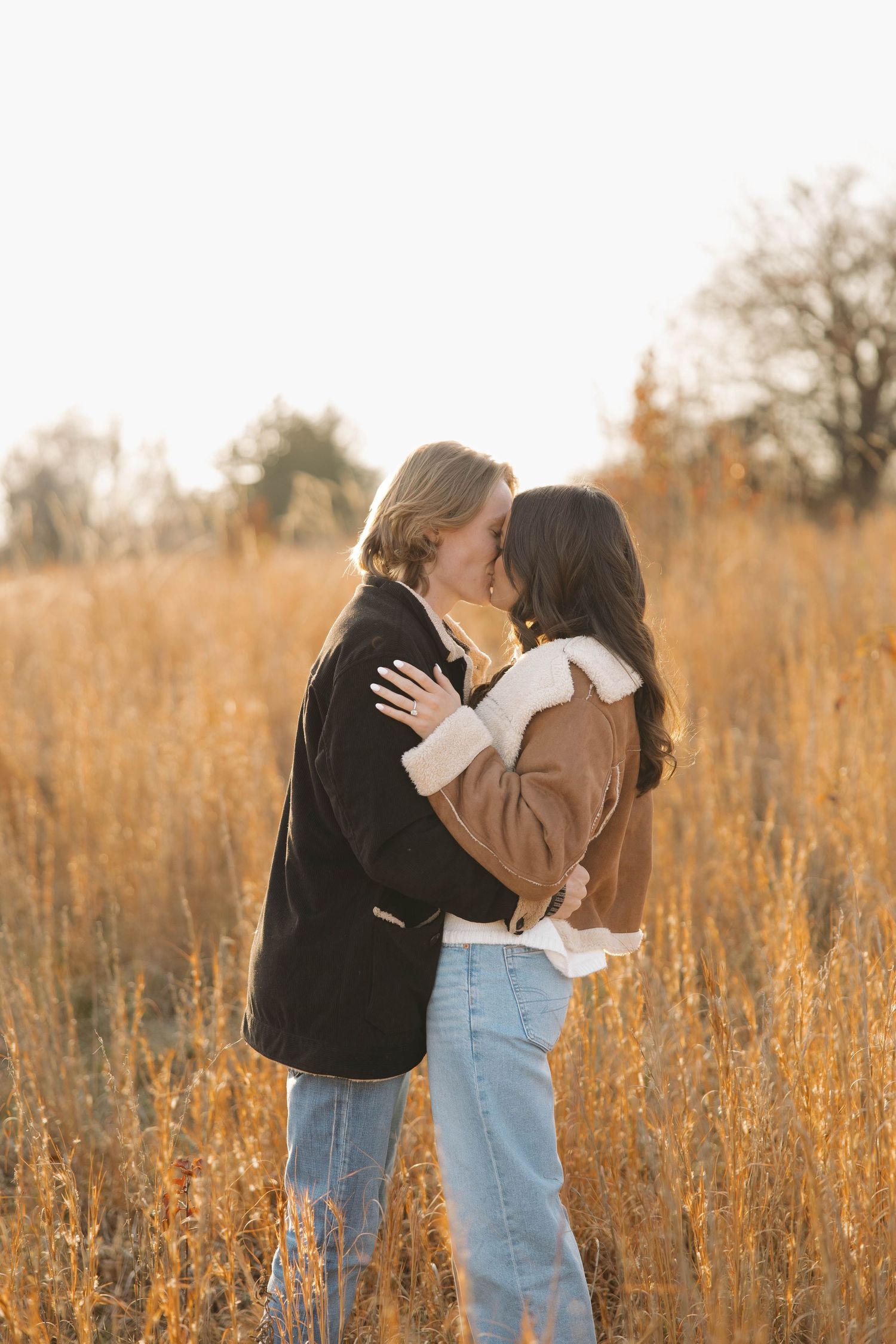 A romantic couple embraces in a golden field during sunset wearing casual outfits of jeans and sweaters.