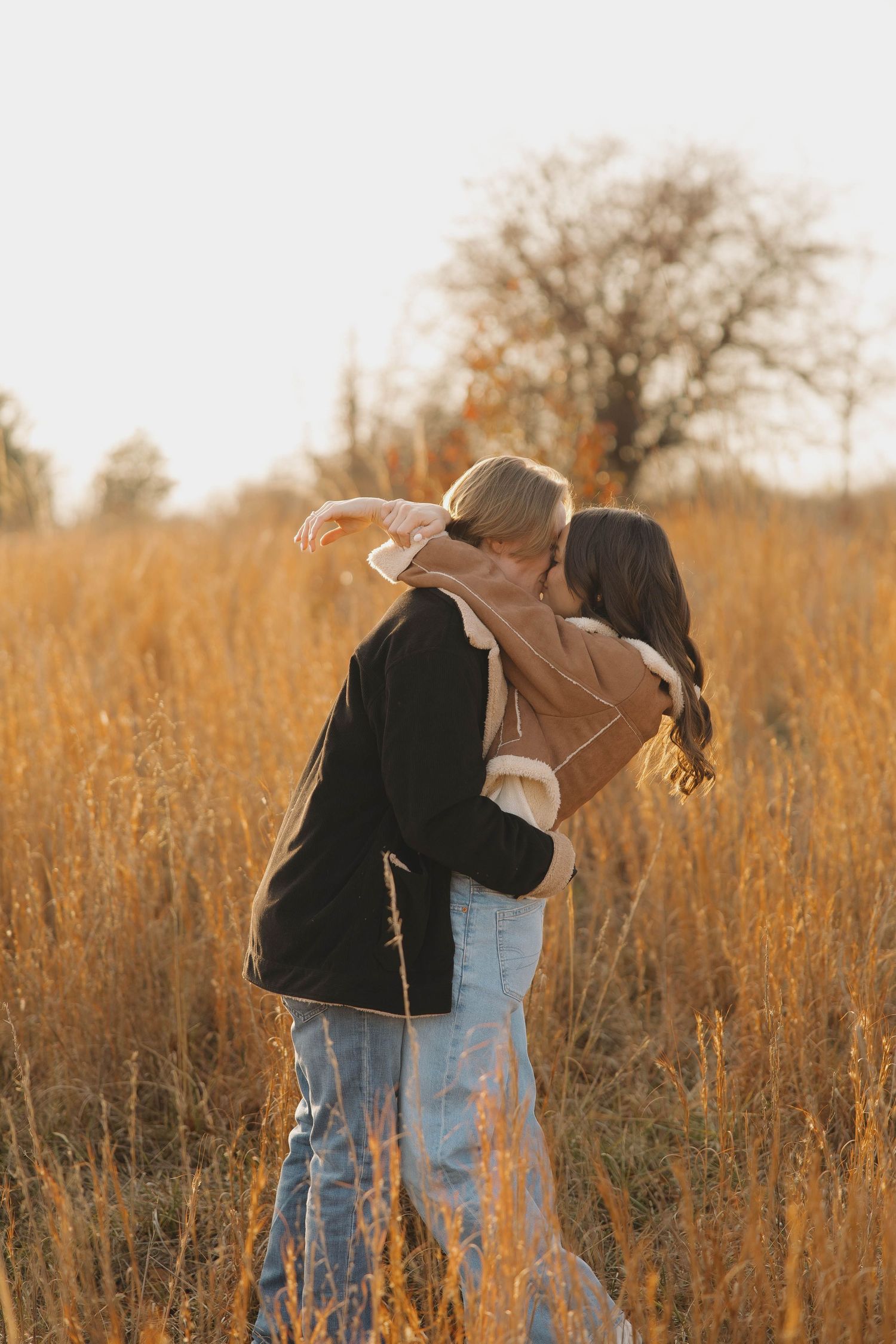 Two people embrace in a golden field of tall grass during sunset with bare trees in the background.