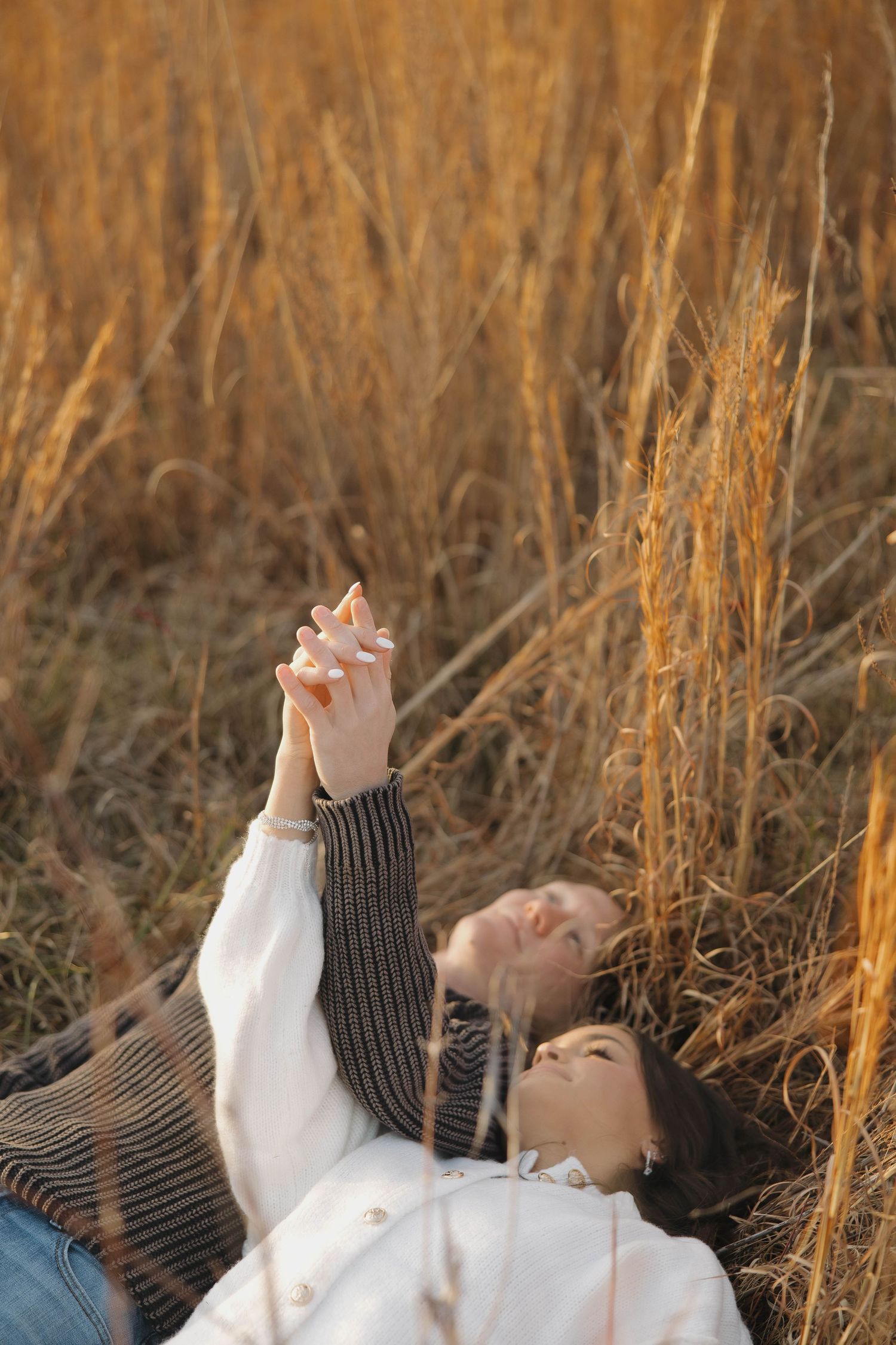 Two people lying in tall grass reaching towards the sky together.