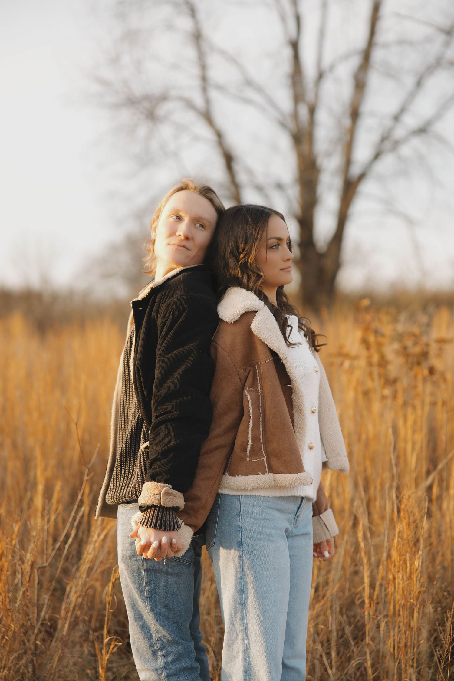 A couple stands back-to-back holding hands in a golden autumn field with bare trees in the background.