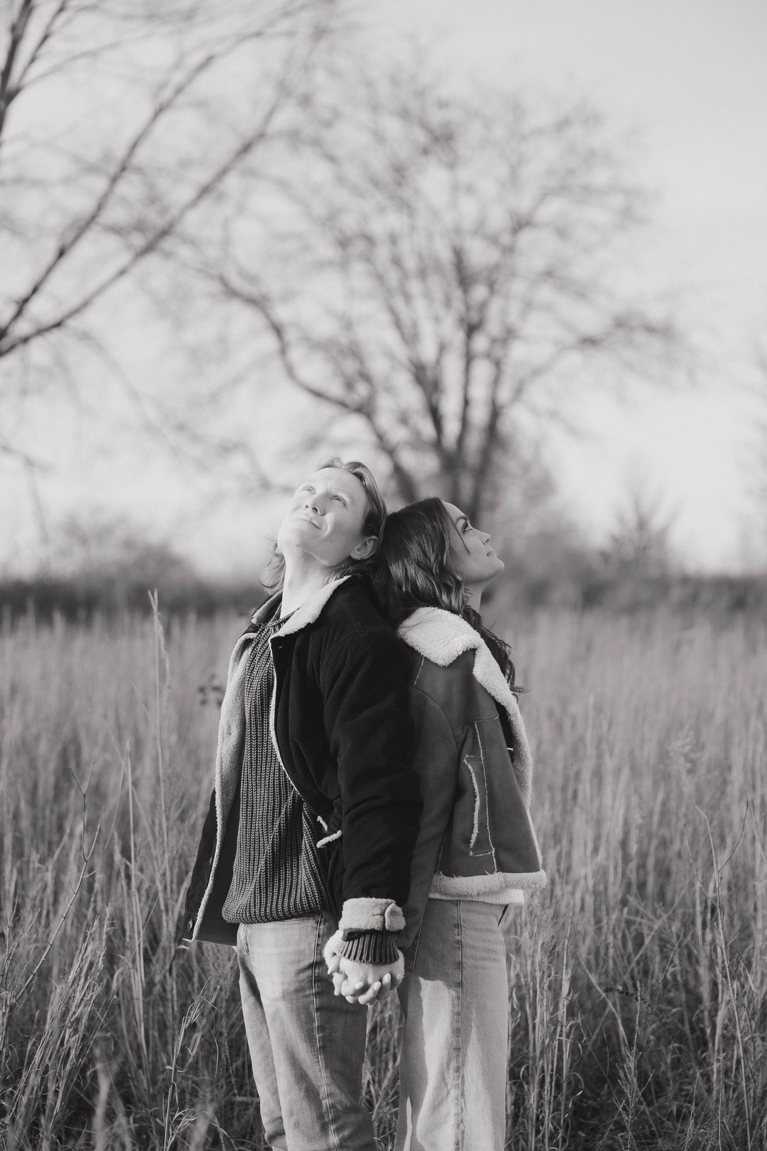 A black and white photo shows two people leaning back-to-back in a field with bare trees in the distance.