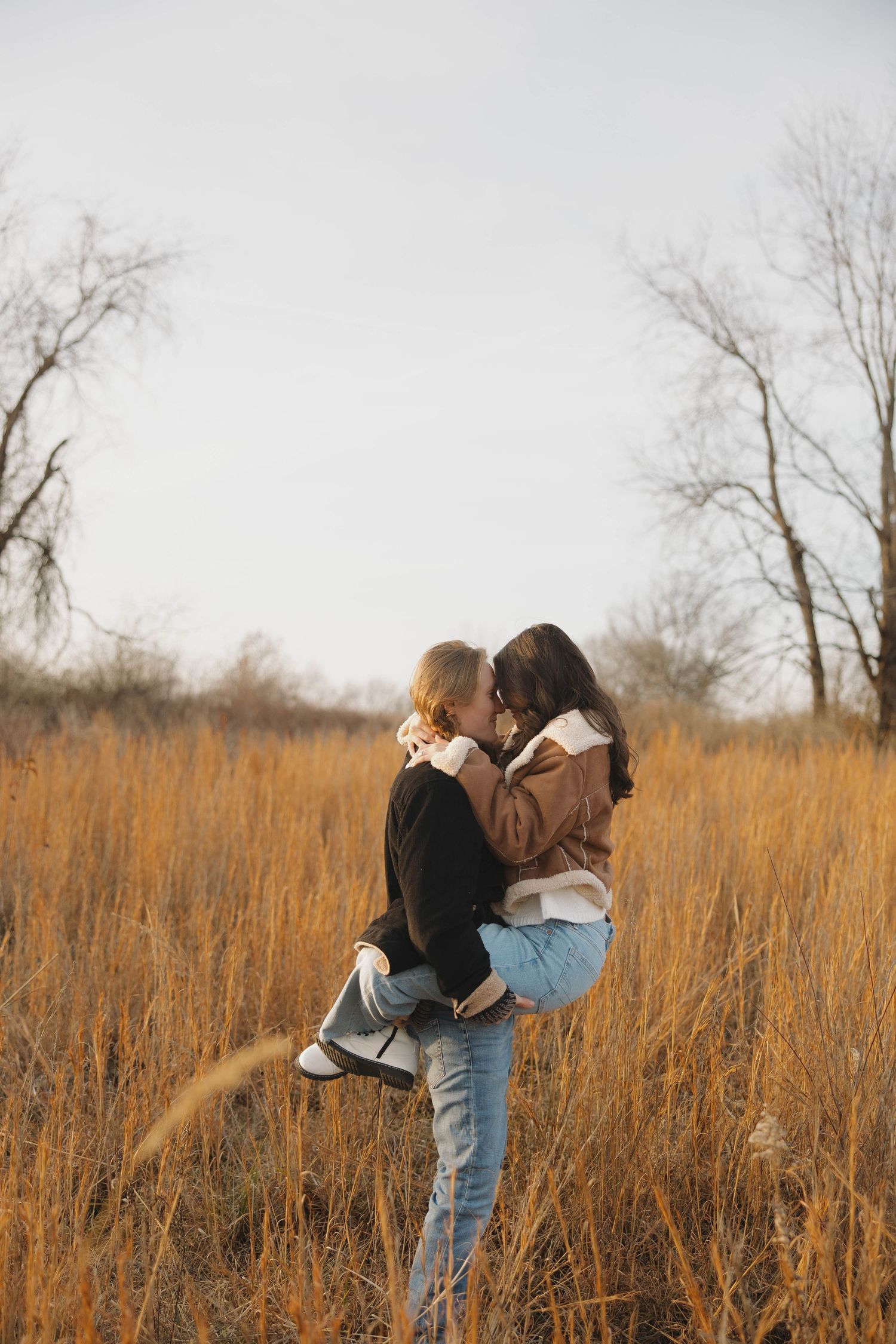 Couple sharing a romantic moment in a golden autumn field with bare trees in background.