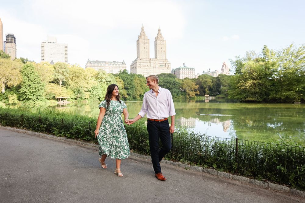 A couple strolls along a path in Central Park with the Manhattan skyline visible in the background.
