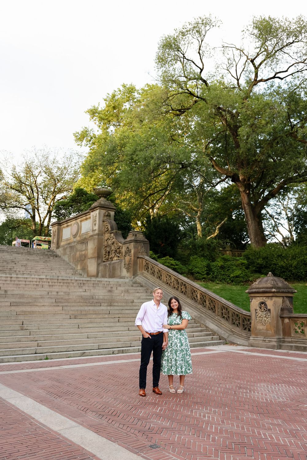 A couple stands on a grand brick terrace with stone steps and ornate railings surrounded by lush trees.