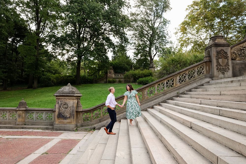 Two people walk down the ornate stone steps at Bethesda Terrace in Central Park on a sunny afternoon.