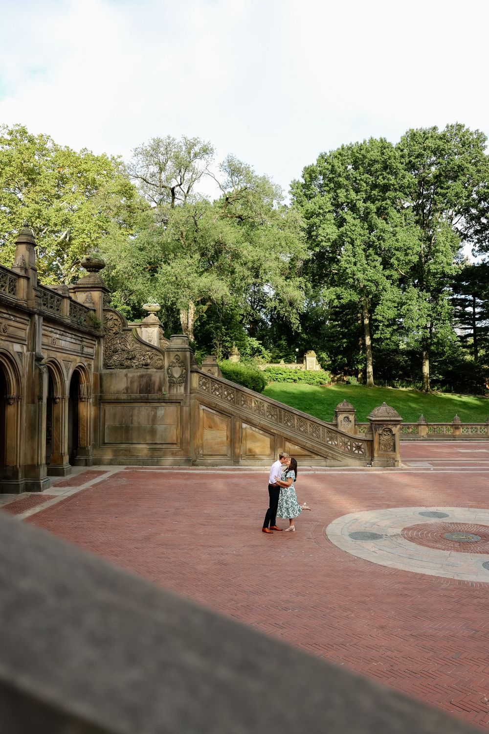 A couple stands together on a red brick terrace surrounded by ornate stone architecture and lush greenery.