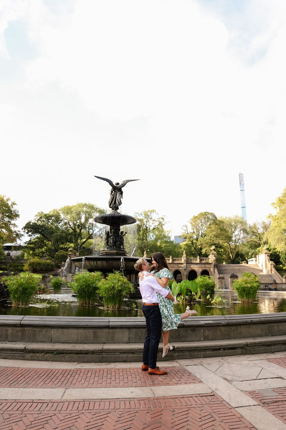 Couple stands near an ornate stone fountain in a park with brick pathways and lush greenery.