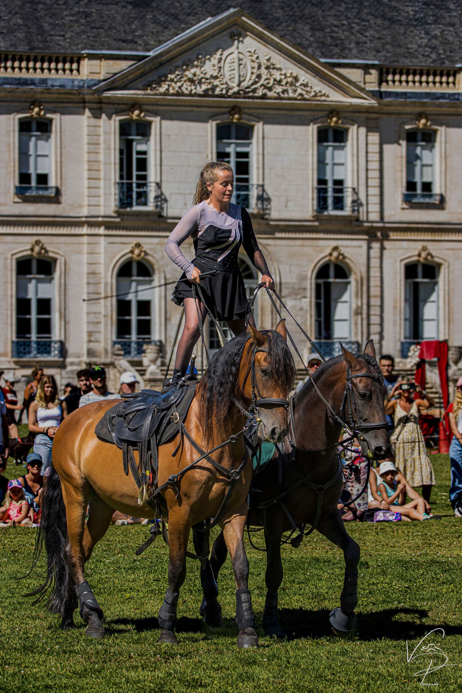 Horses and riders perform on grass in front of a grand historic building with spectators watching.