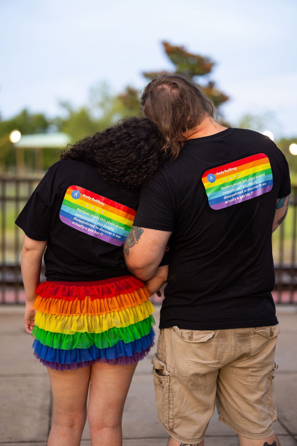Couple wearing matching black rainbow pride t-shirts, seen from behind, one in a rainbow tutu skirt.