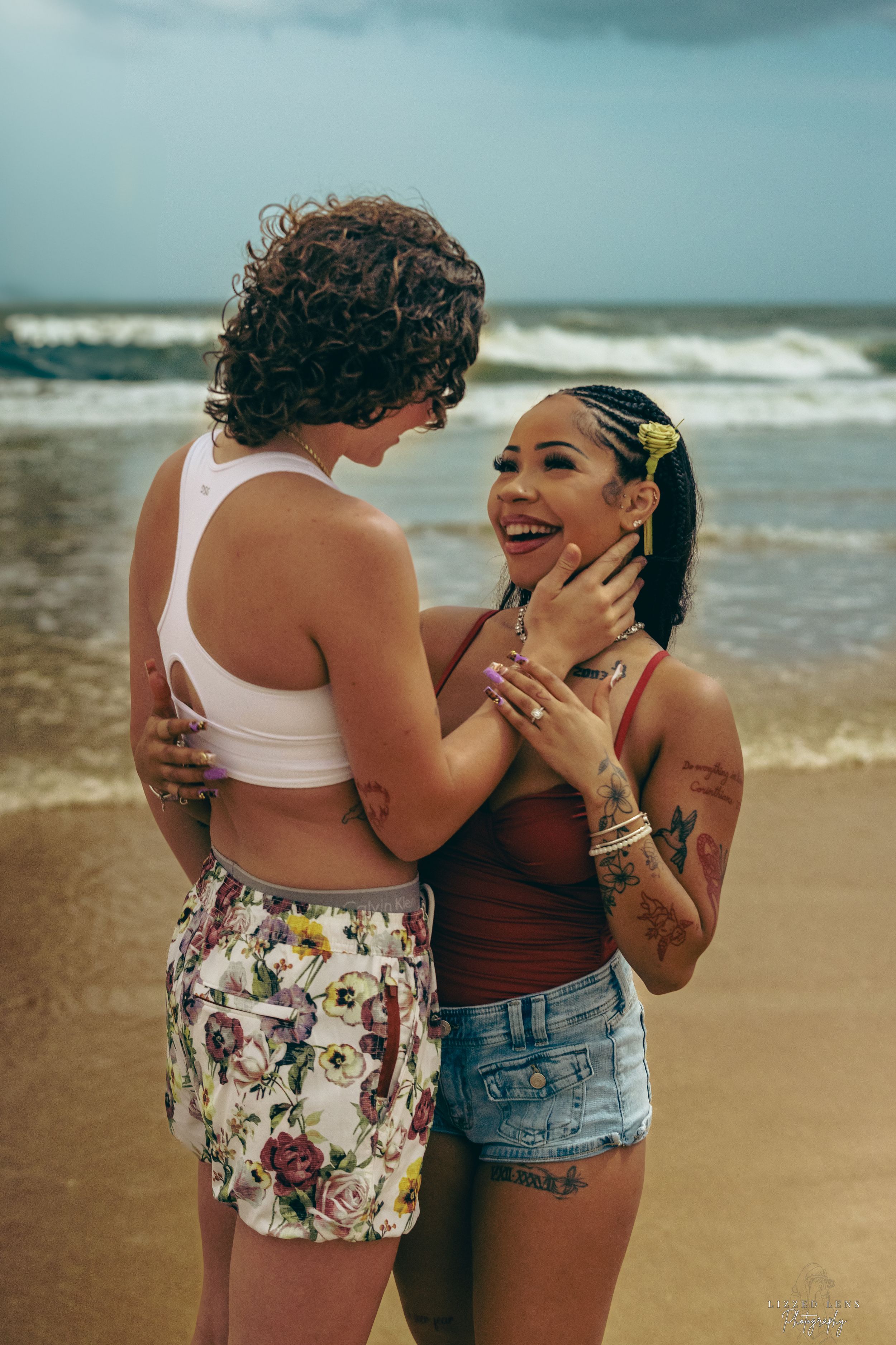 Two tattooed women laugh and embrace joyfully on a sandy beach with ocean waves behind them.