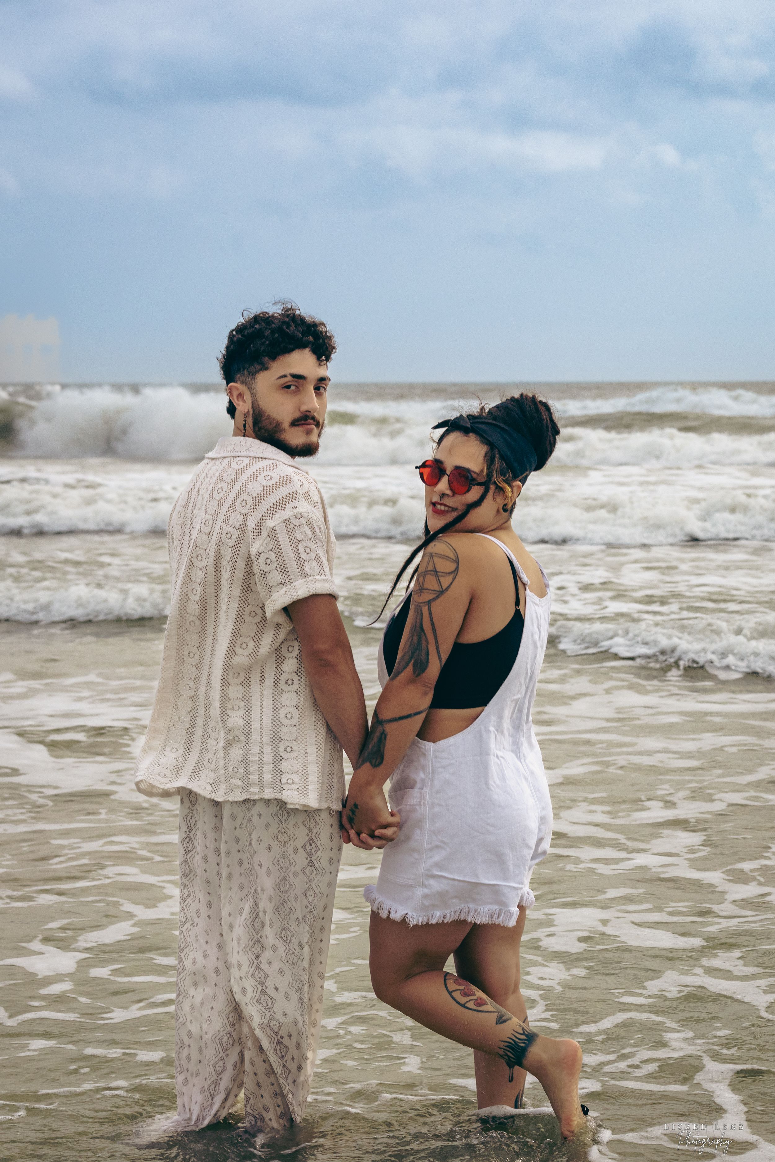 A couple holds hands and looks back while standing in ocean waves on a cloudy beach day.
