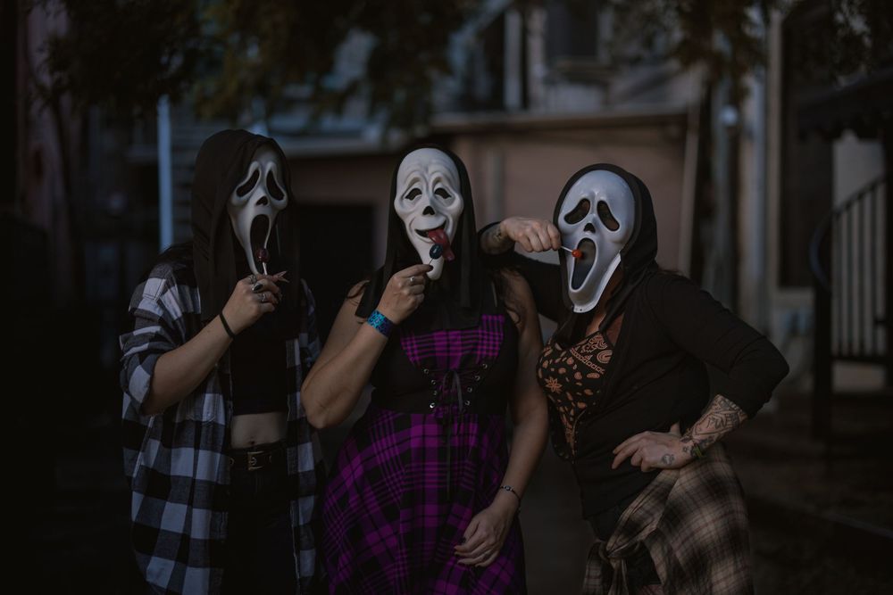 Three friends in plaid and dark clothing pose outdoors in Ghostface-style Halloween masks, striking playful spooky poses at dusk.