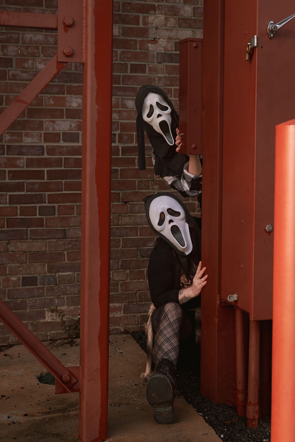 Two people in Ghostface-style masks and black clothing peek around red metal beams against a brick wall in a spooky Halloween scene.