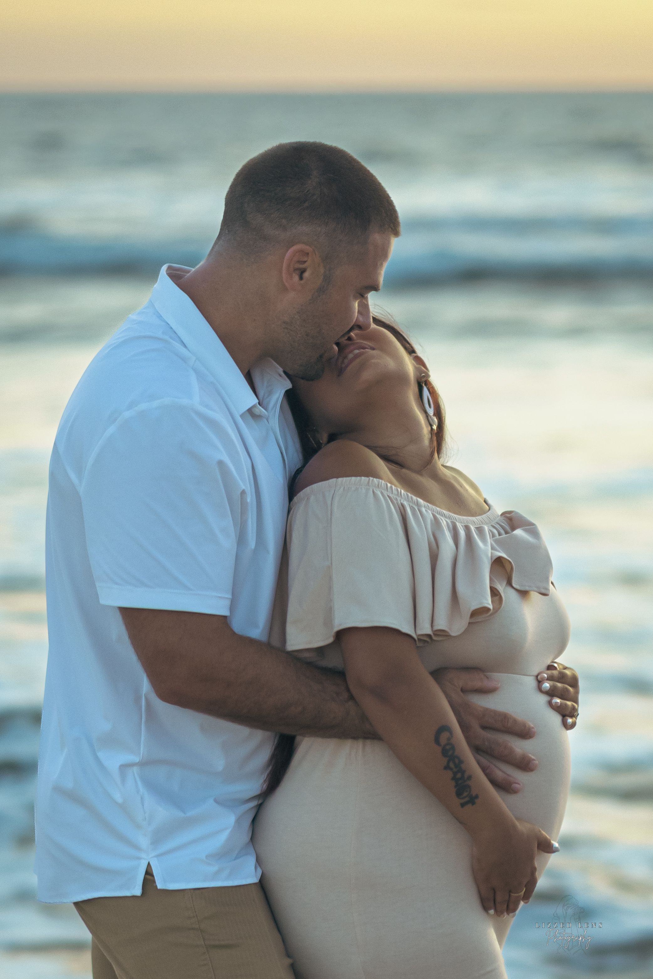 A couple shares a tender kiss on the beach at sunset while the woman cradles her pregnant belly.