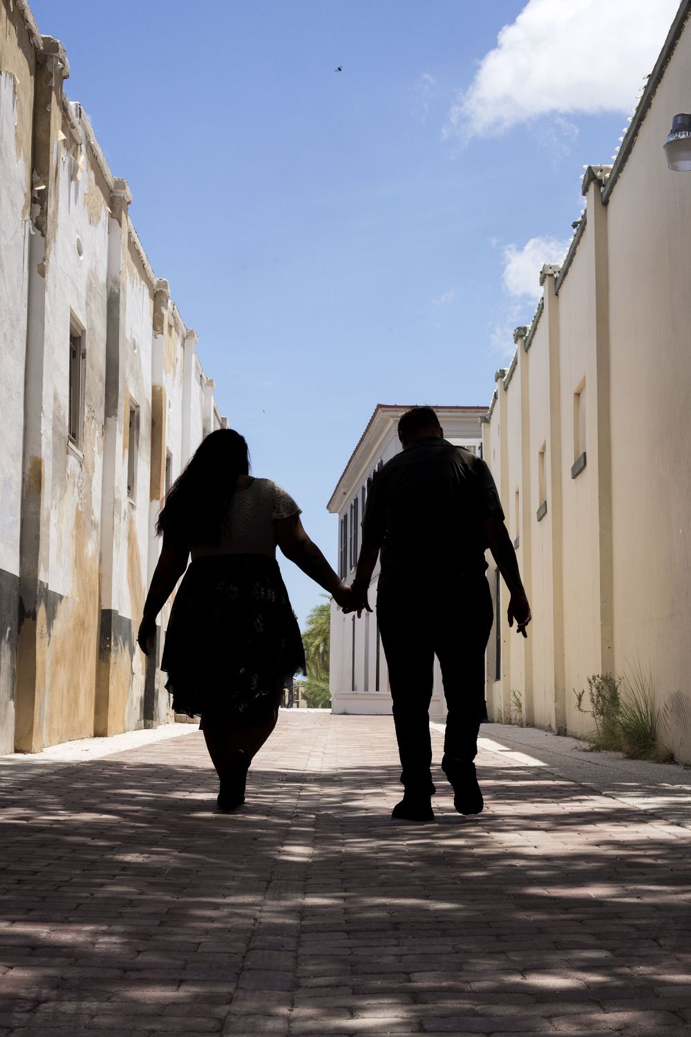 Silhouette of a couple holding hands walking down a sunlit brick pathway between historic buildings under blue sky.