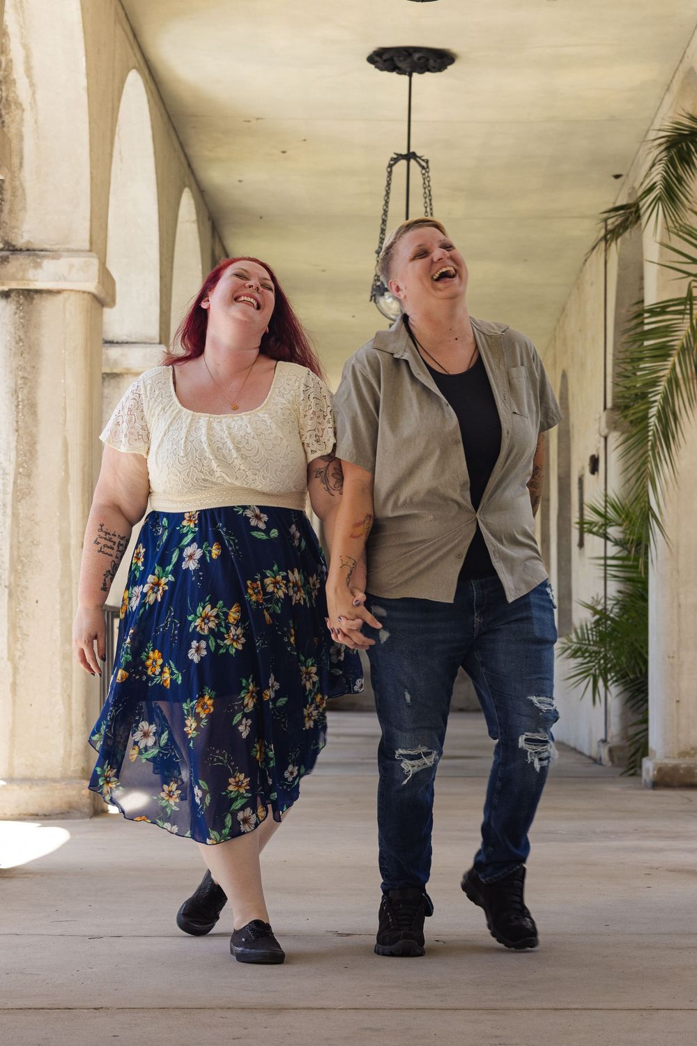 Two women holding hands and laughing while walking together through an arched outdoor corridor.