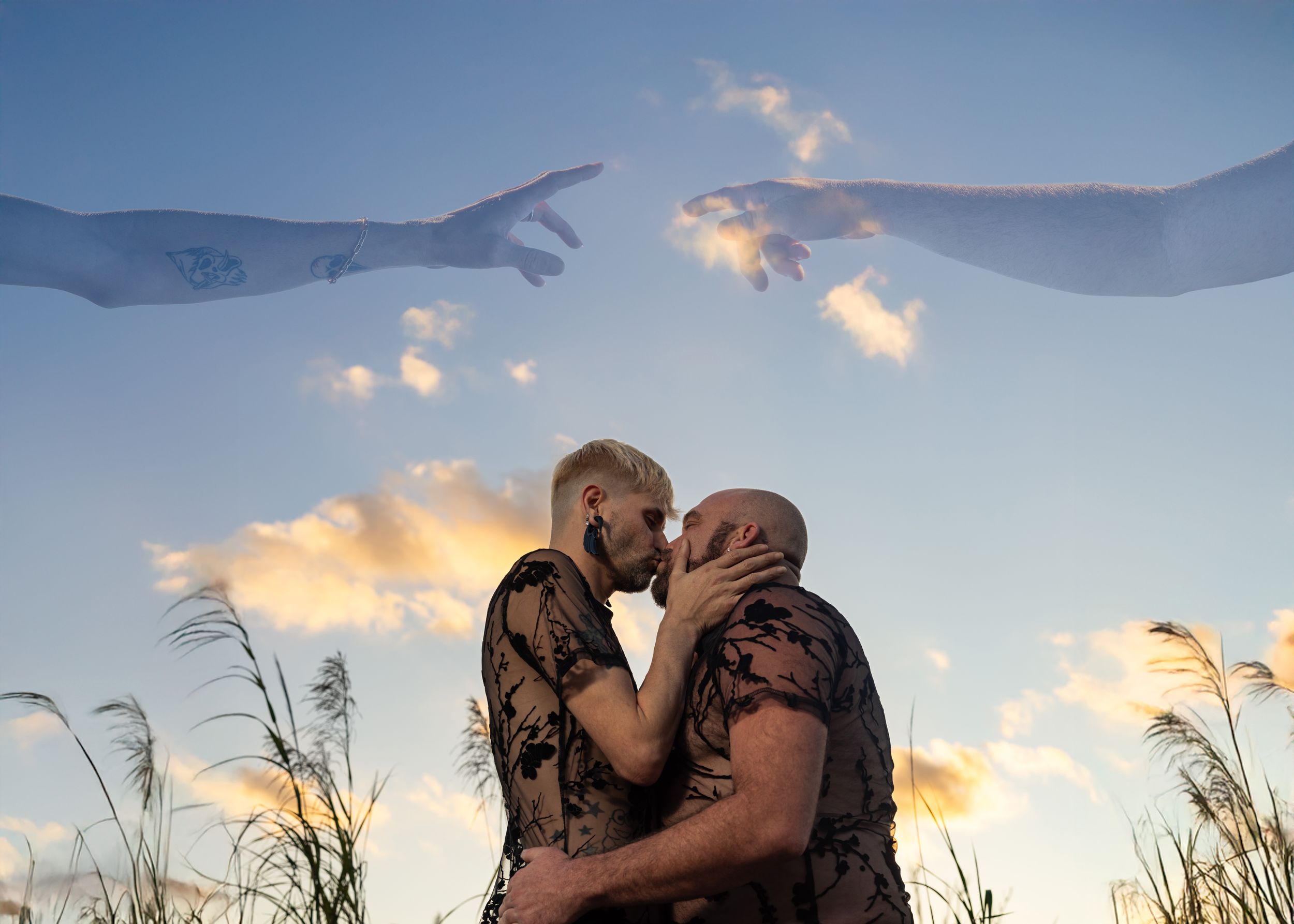 Two men share a romantic kiss outdoors with artistic hands reaching from clouds above in a dreamy sky.