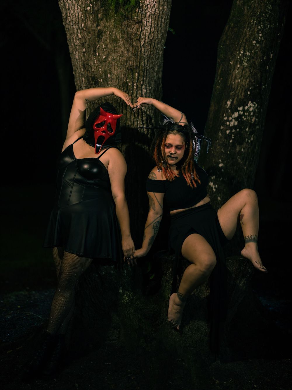 Two women in dark gothic outfits pose among tree trunks at night, one wearing a red devil mask and arms raised in a heart shape.