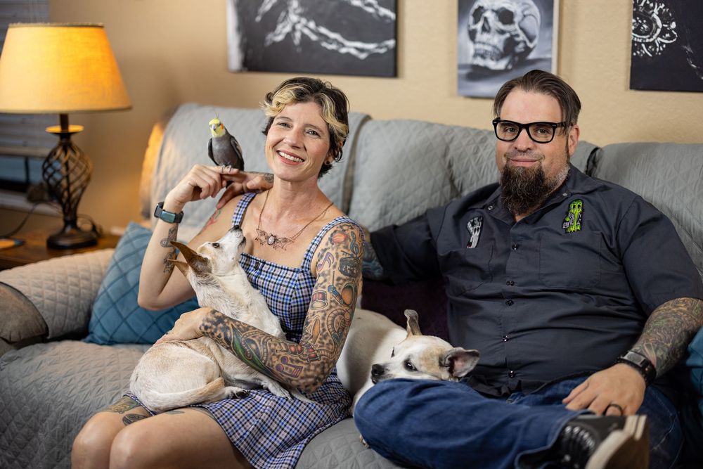 A tattooed couple sits on a couch with a small dog and a cockatiel bird perched on the womans hand.