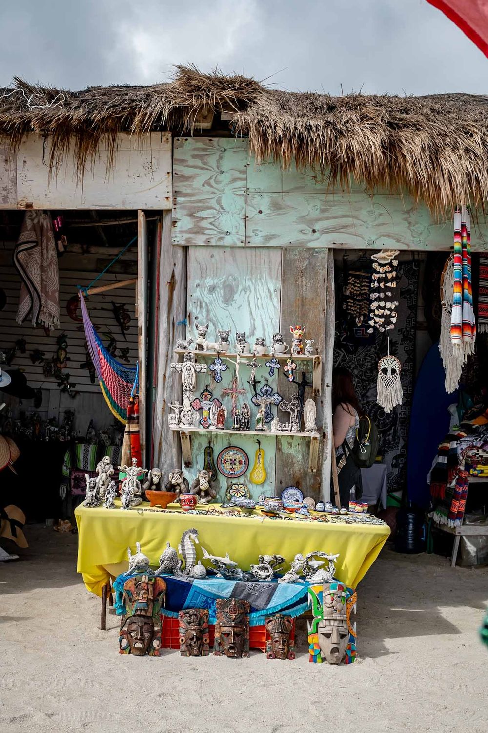 Rustic beachside market stalls with thatched roofs display colorful handmade crafts and souvenirs for sale.