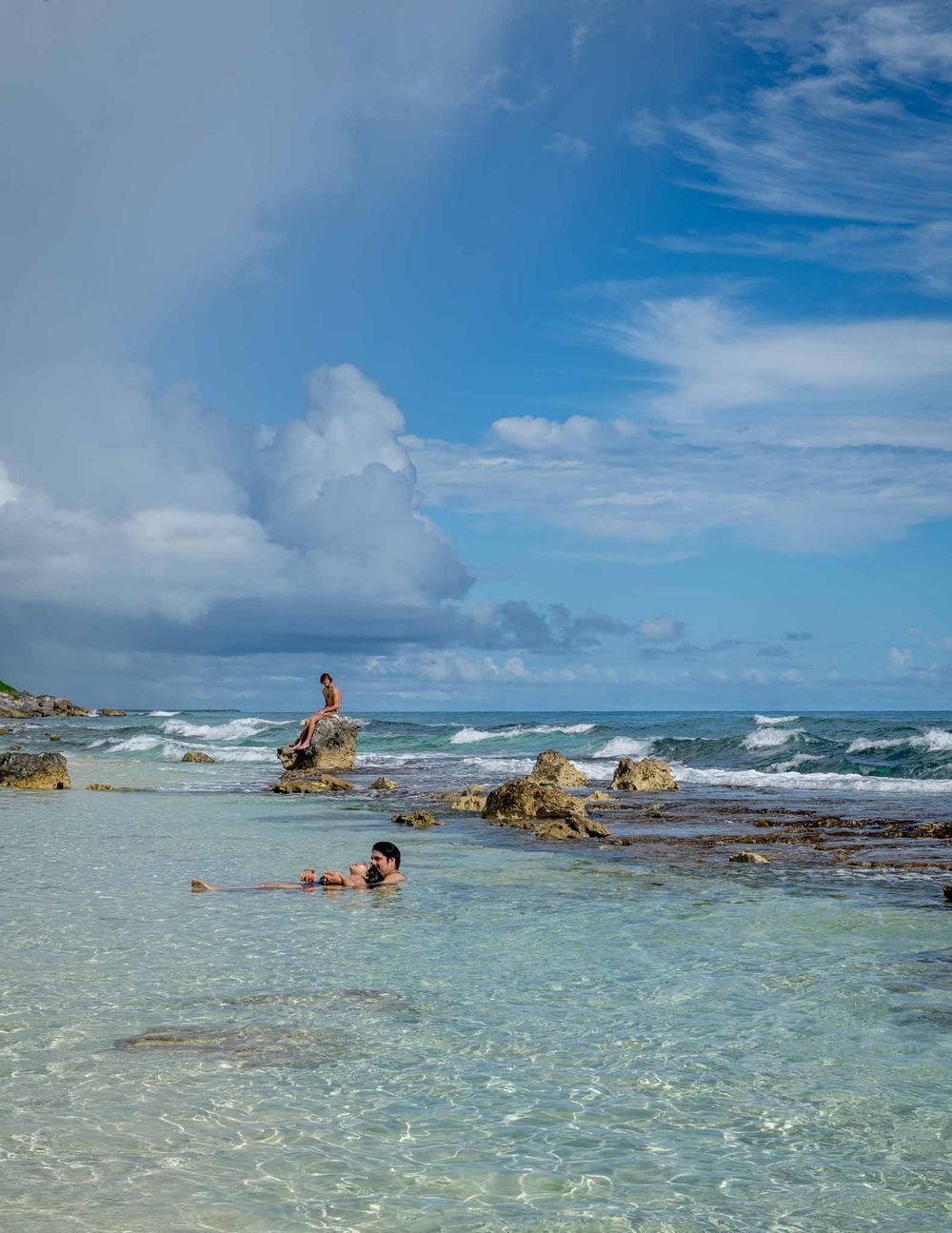 Panoramic view of a crystal clear tropical beach with turquoise waters and rocky outcrops under a bright blue sky.