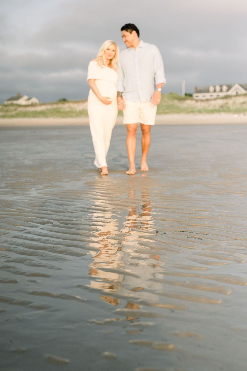 Two people in white clothing walking along a sandy beach at low tide with their reflections visible in the wet sand.