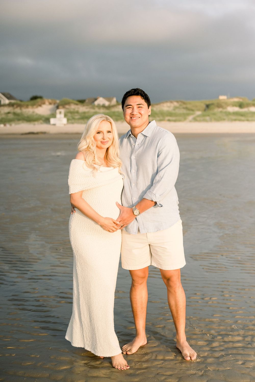 A couple poses together on a sandy beach at sunset wearing white and beige clothing.