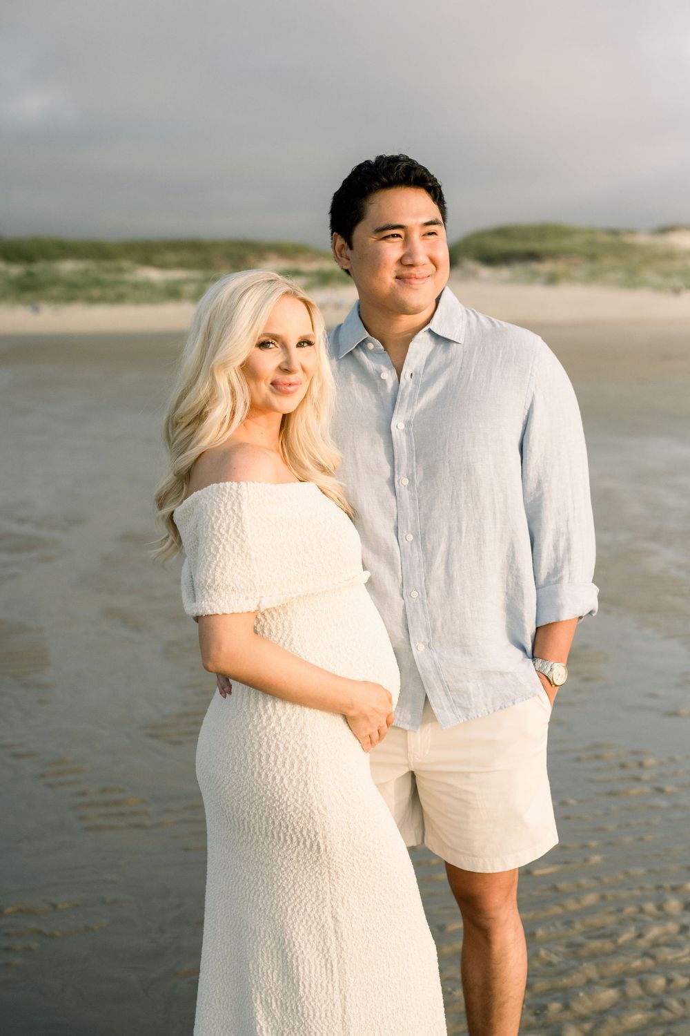 A couple in white attire poses together for romantic maternity photos on a sandy beach at sunset.