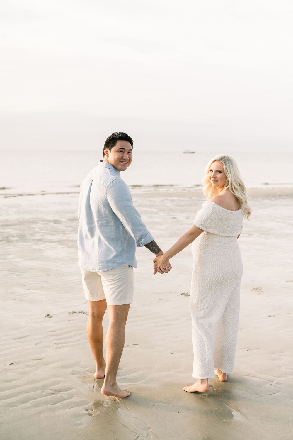 A couple in white attire walks hand in hand along a serene beach during a romantic photoshoot.