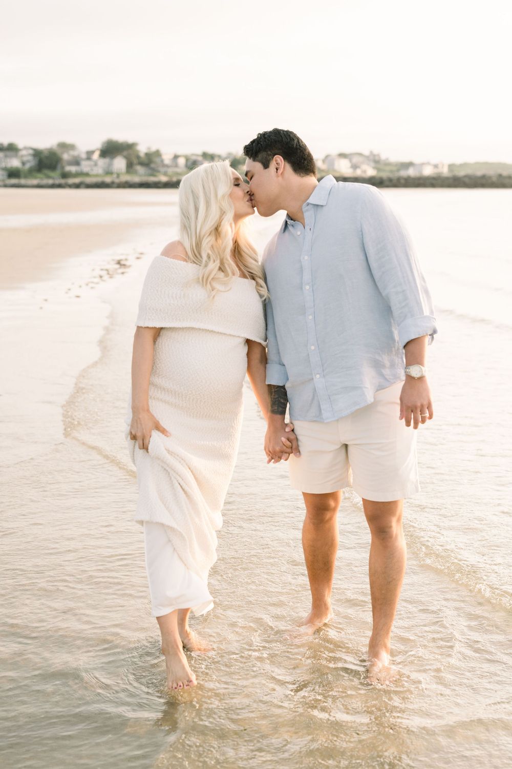 A romantic couple shares a tender moment while standing barefoot in shallow beach water at sunset.